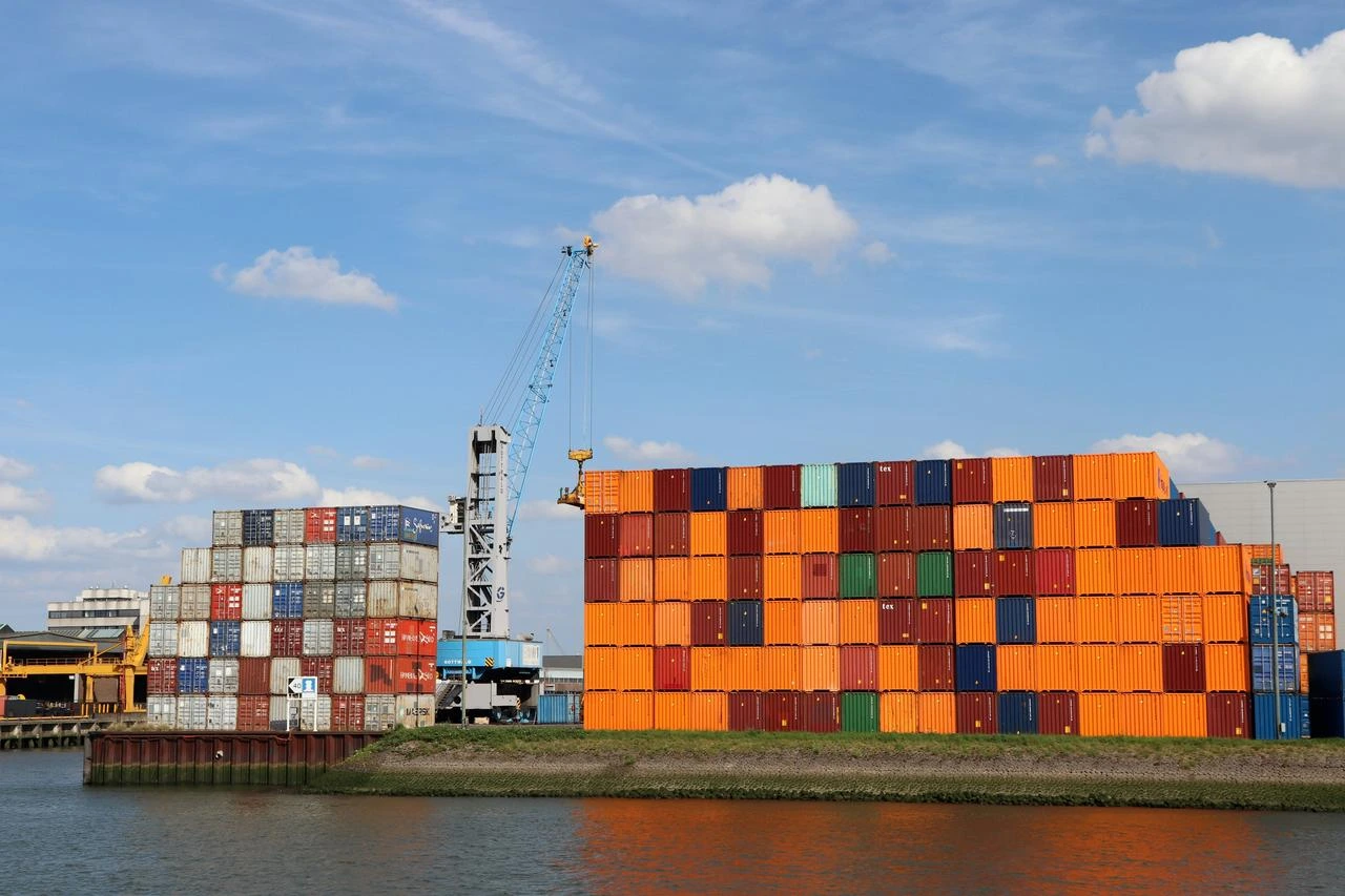 Container terminal with stacked shipping containers, cranes, and port infrastructure under a clear blue sky.